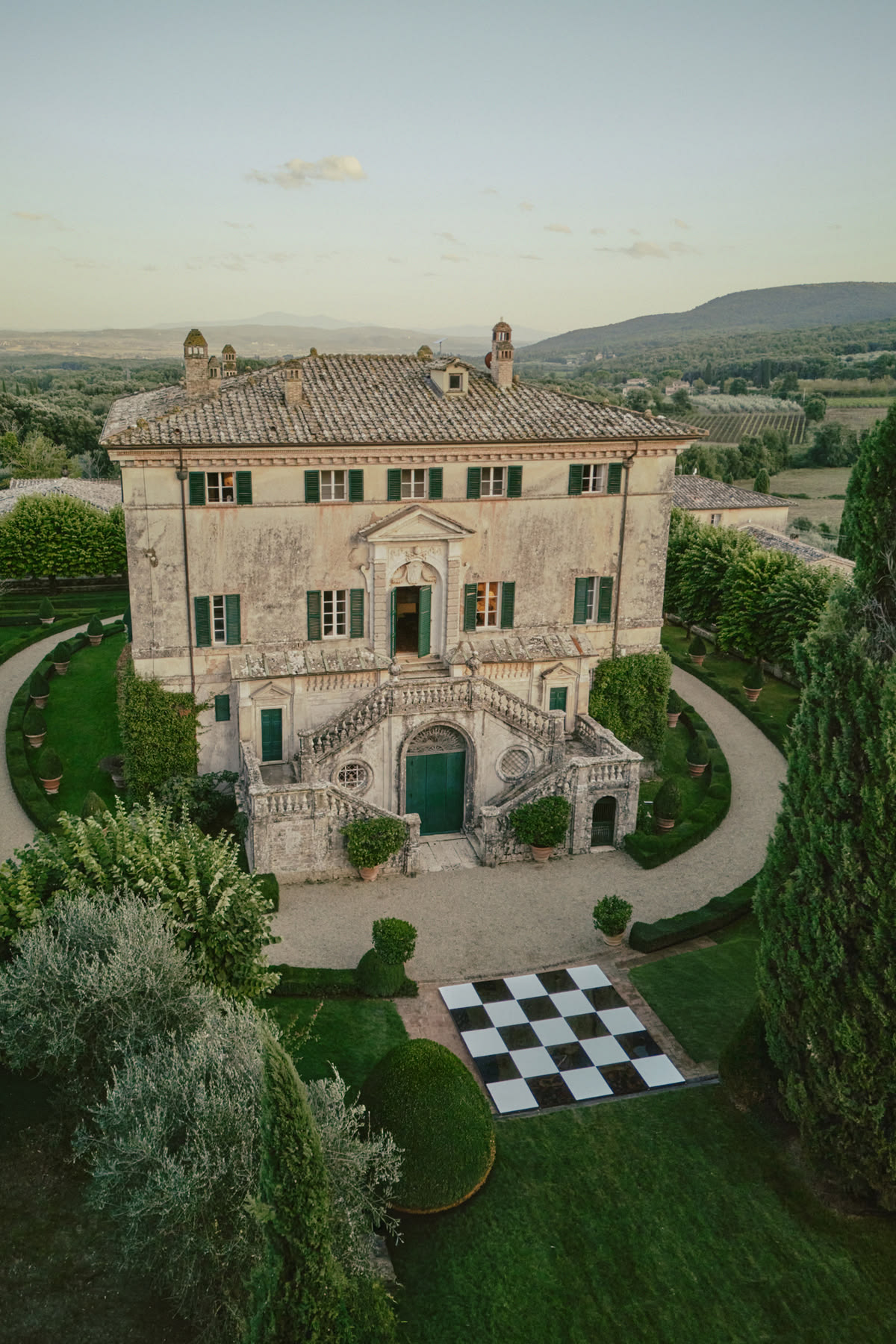 Elevated view of a historic stone villa with green shutters and a terracotta roof, a black-and-white checkered dance floor laid out on the lawn, formal gardens and rolling hills beyond