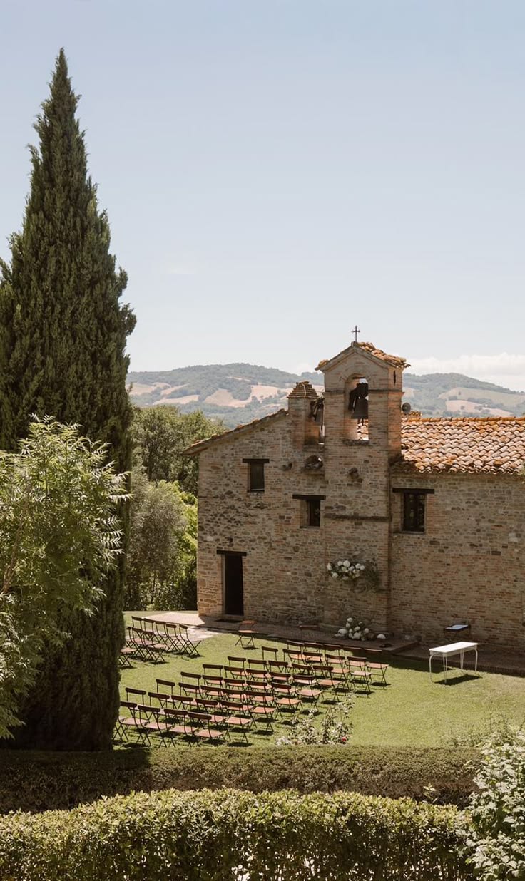 A rustic stone country chapel with a bell gable and cross, rows of wooden ceremony chairs arranged on the lawn in front, cypress trees and rolling hills behind